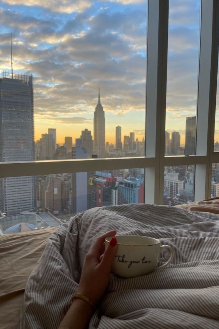 a woman laying in bed with a cup on her lap and looking out the window