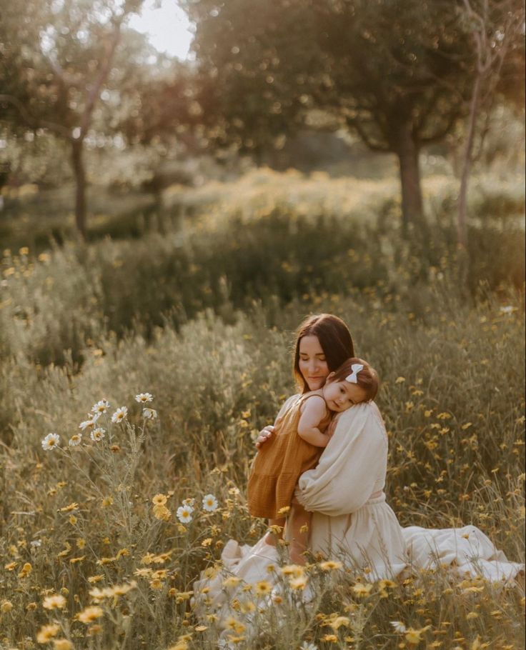 a woman holding a baby in a field full of wildflowers and trees at sunset