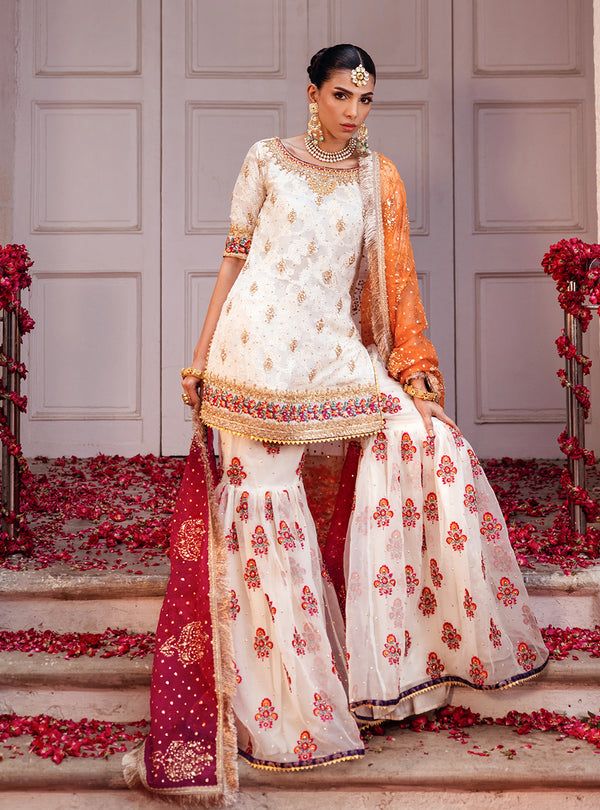 Embrace the essence of tradition and elegance with our stunning Chunri chiffon off-white ensemble, adorned with intricate machine work and heavy hand embroidery on the neck, hem, and sleeves. Paired perfectly with an organza embroidered gharara, accentuated with light handwork and scattered chan, creating a harmonious blend of texture and elegance. The look is completed with a gradient organza dye block print dupatta, embellished with sequin and gota spray and the finishing of jamwar and kiran a Designer White Sharara With Dupatta, Festive White Raw Silk Palazzo Set, White Bollywood Style Raw Silk Palazzo Set, Designer White Raw Silk Palazzo Set, White Raw Silk Palazzo Set For Designer Wear, Designer White Palazzo Set With Intricate Embroidery, Off White Georgette Palazzo Set With Dupatta, White Silk Traditional Designer Wear, Designer White Silk Traditional Wear