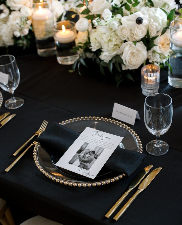 a black table cloth with white flowers and candles on it is set for a formal dinner