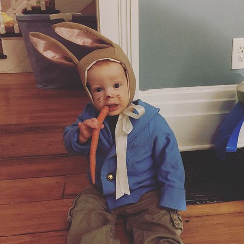 a little boy sitting on the floor wearing a bunny ears hat and holding a carrot in his mouth