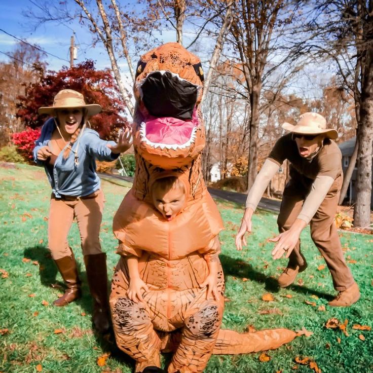 two people in costumes playing with an inflatable dinosaur on the grass near trees