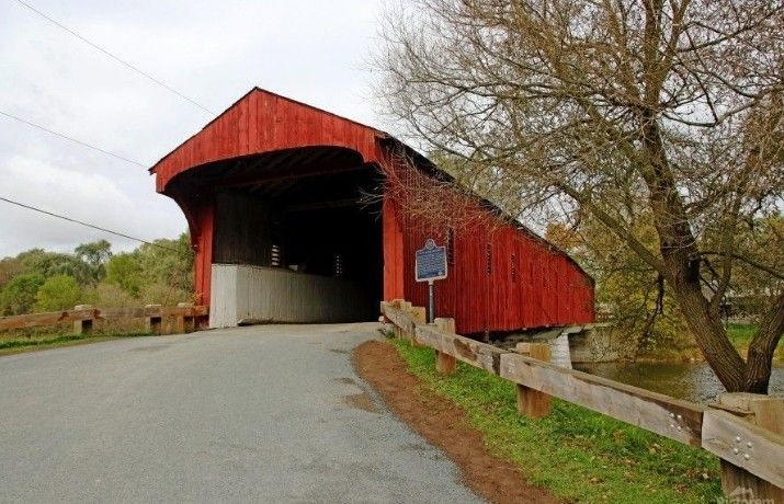 a large red covered bridge over a small river next to a tree with no leaves on it