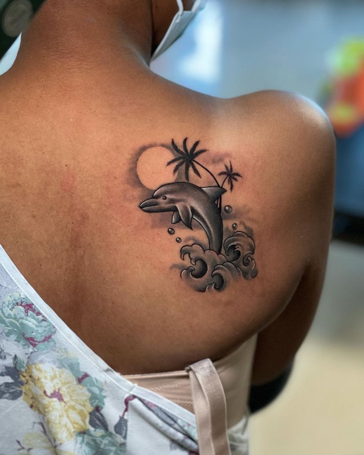 a woman's back with a dolphin tattoo on her left shoulder and palm trees in the background