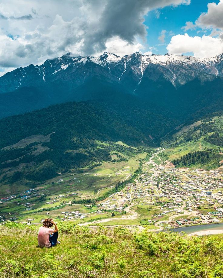 a man sitting on top of a lush green hillside next to a valley filled with mountains