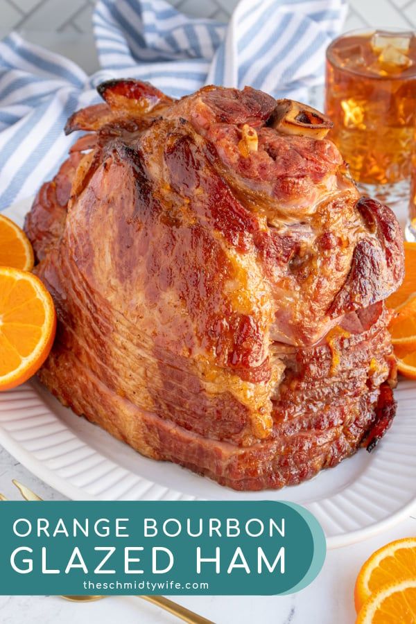 an orange bourbon glazed ham on a white plate next to sliced oranges and two glasses