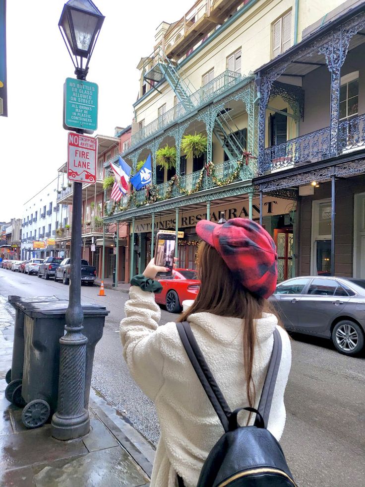 a woman is standing on the sidewalk taking a photo with her cell phone while wearing a red and black hat