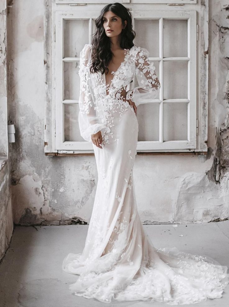 a woman standing in front of a window wearing a wedding dress with long sleeves and an open back