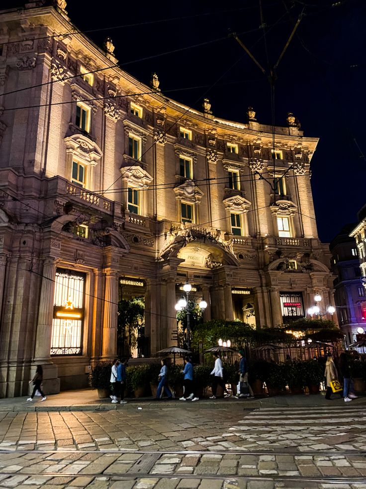 people are walking around in front of an old building at night with the lights on