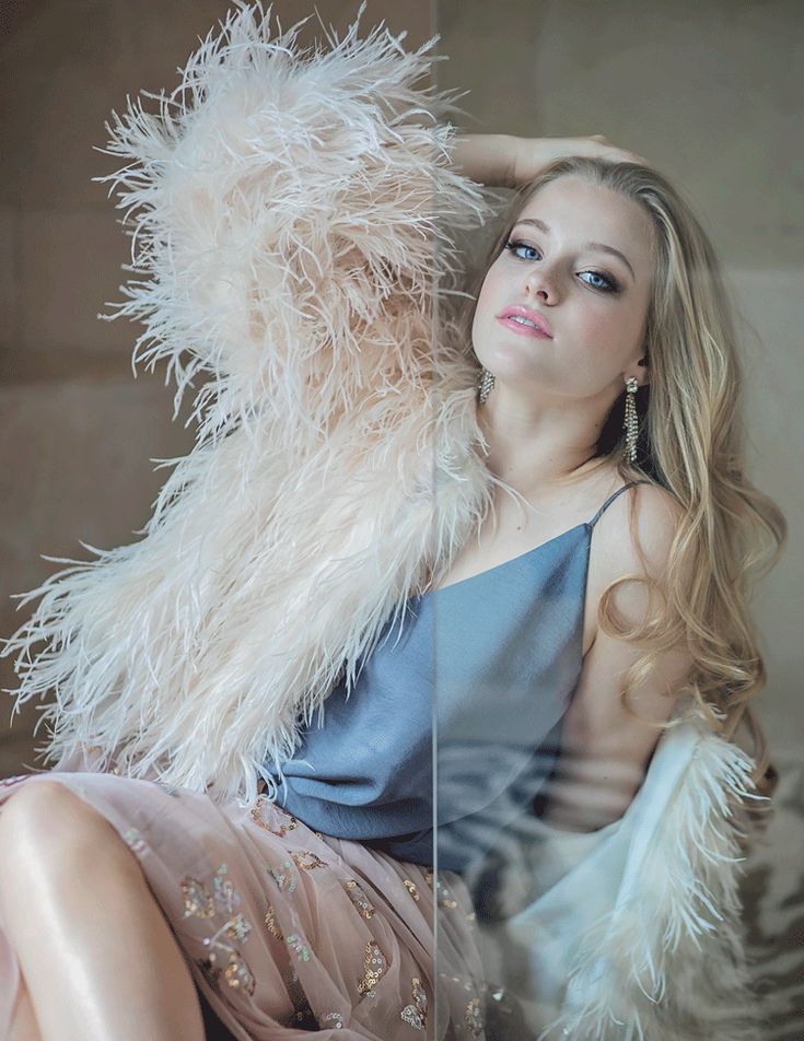 a woman in a dress and feather jacket sitting on a couch with her arms behind her head