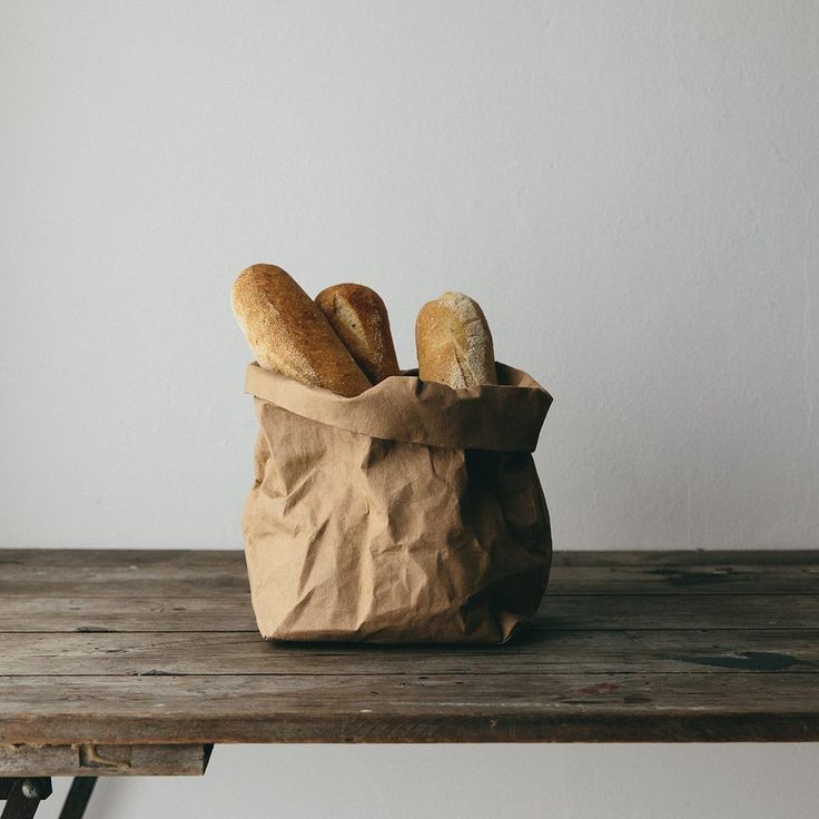 two loaves of bread in a brown paper bag on a wooden table next to a white wall