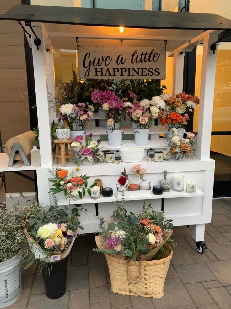 some flowers are sitting on top of a white stand with a sign that says give a little happiness