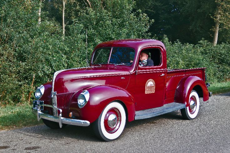 an old red truck is driving down the road in front of some bushes and trees