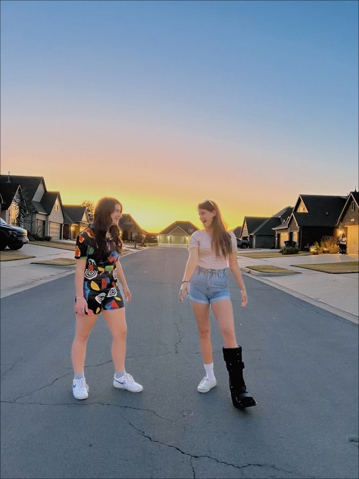 two young women standing in the middle of an empty street at sunset with houses in the background