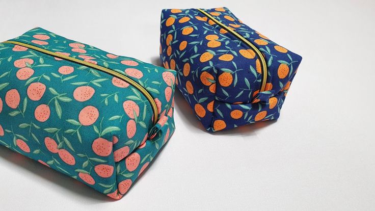 two cosmetic bags sitting next to each other on a white surface with oranges and green leaves