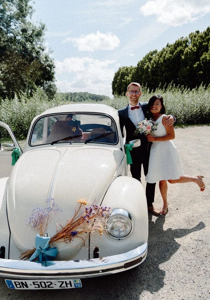 a man and woman standing next to an old car with flowers in the front seat