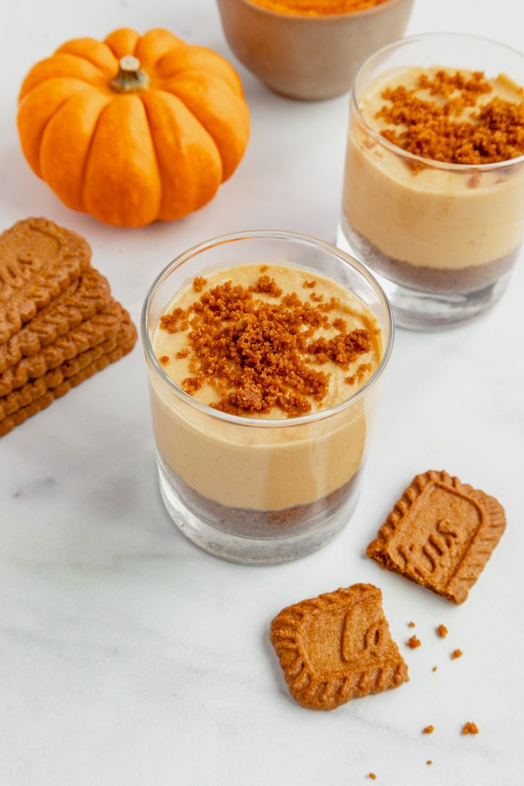 two glasses filled with desserts next to cookies and pumpkins on a white table