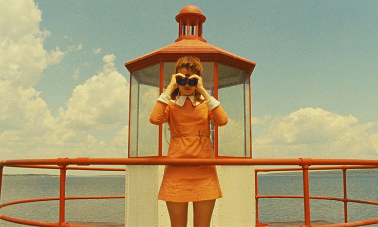 a woman in an orange dress standing on a boat looking at the ocean through binoculars