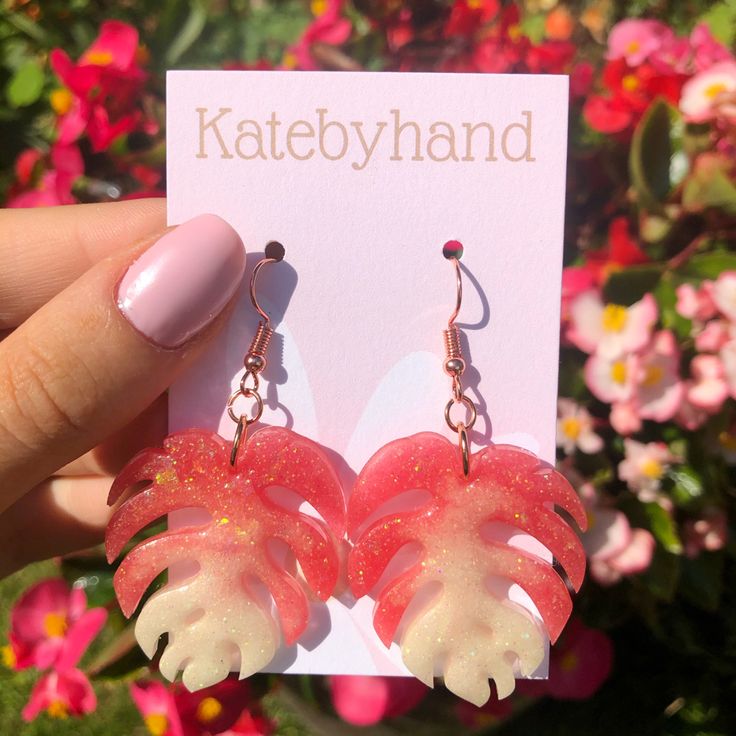 a pair of pink and white flower shaped earrings with dangling earwires in front of flowers