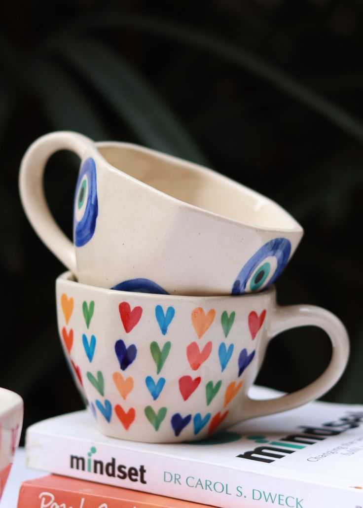 two cups with hearts painted on them are sitting next to some books and coffee mugs