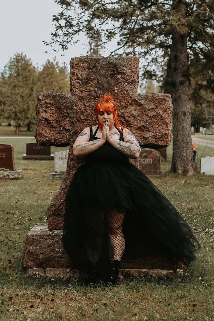 a woman with red hair wearing a black dress sitting on a rock in a cemetery