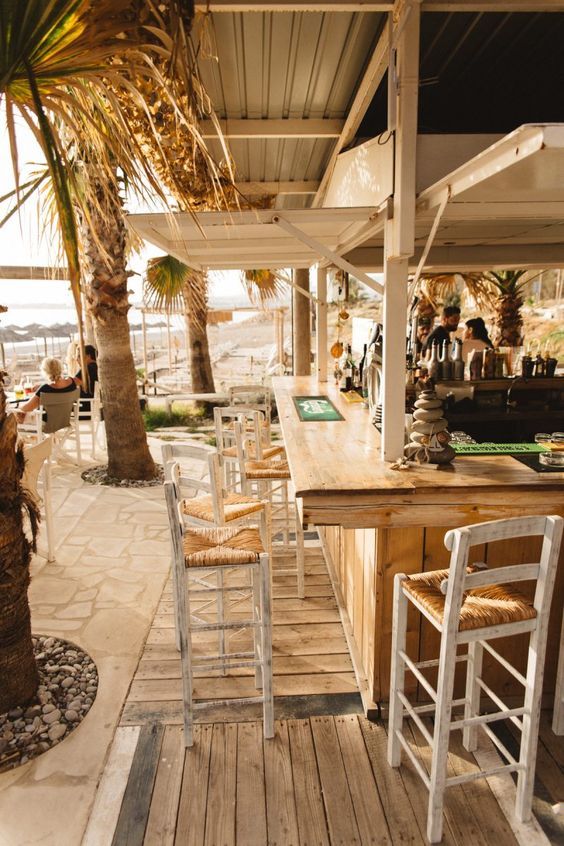 an outdoor bar with wooden tables and chairs next to palm trees on the beach side