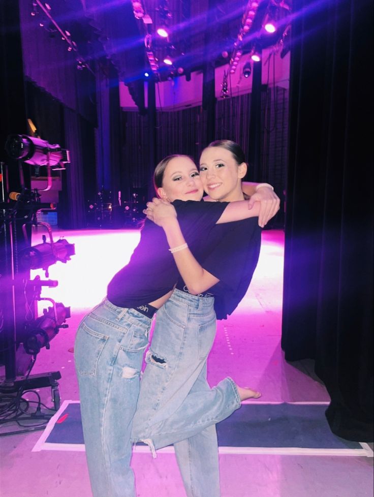 two women hugging each other in front of a stage with lights on the floor and purple lighting behind them