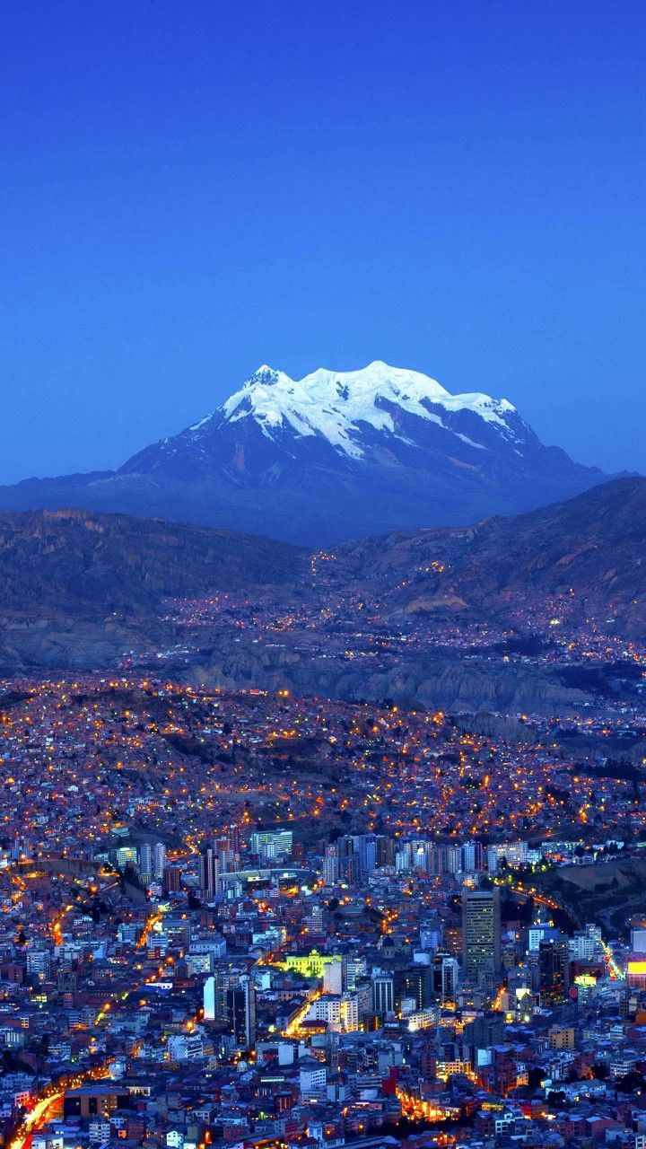 a city with snow capped mountains in the background at night time, lit up by street lights