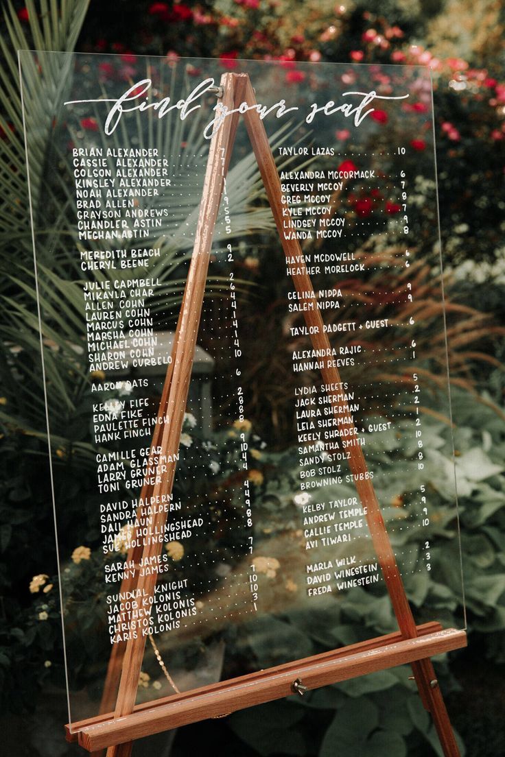 a wooden easel sitting in front of a flower garden with an acrylic sign on it