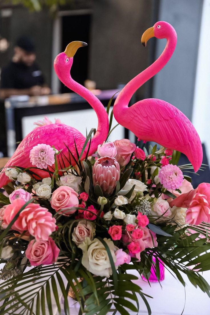 two pink flamingos sitting on top of a vase filled with flowers and greenery