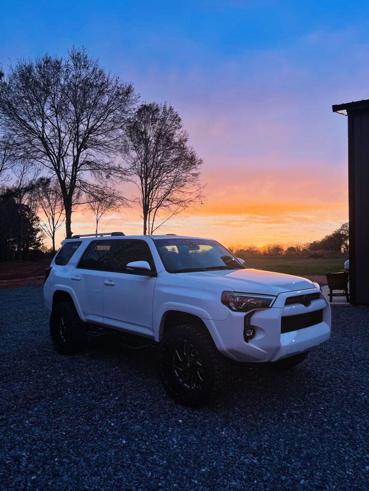 a white toyota 4runner is parked in the parking lot at sunset with trees behind it