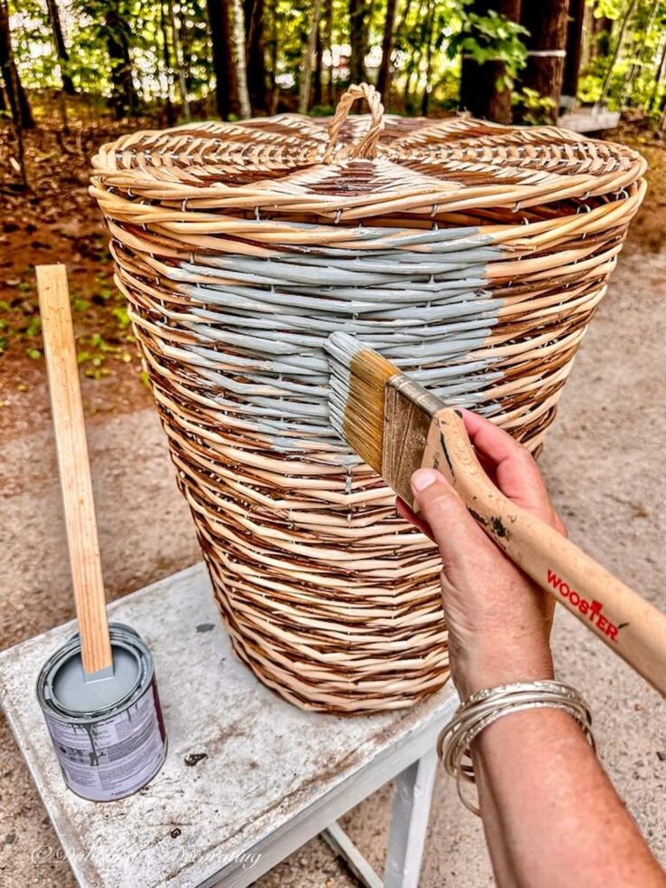 a person painting a basket with paintbrushes on a table in front of some trees