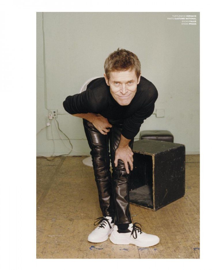 a man in black shirt and leather pants leaning on an old suitcase with white shoes