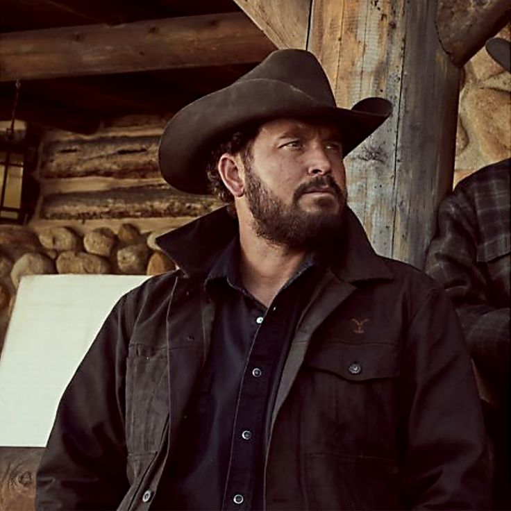 a man with a beard wearing a cowboy hat and black shirt standing in front of a log cabin