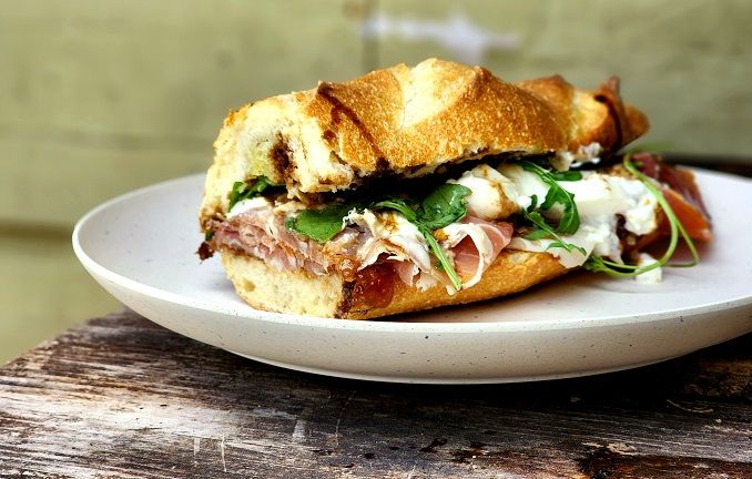 a sandwich with meat, cheese and greens on a white plate sitting on a wooden table