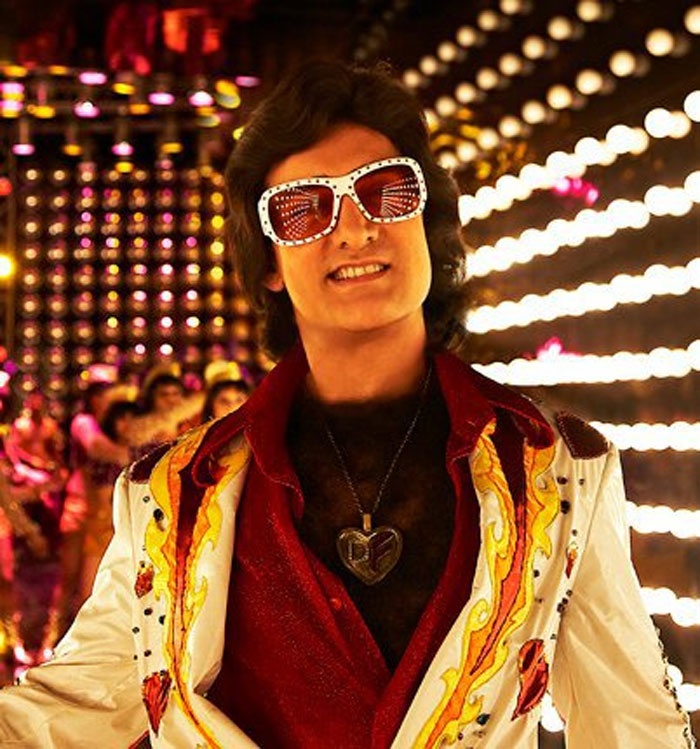 a man with sunglasses on standing in front of some people wearing costumes and lights behind him