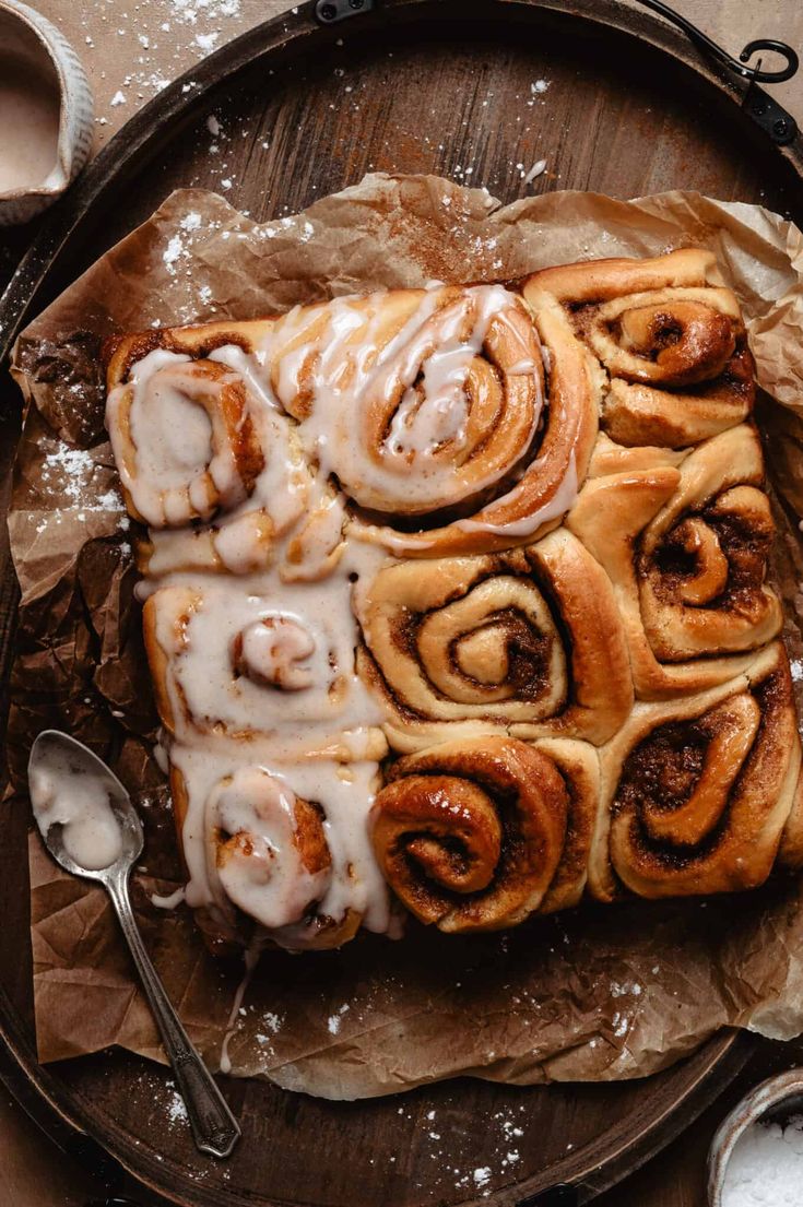 cinnamon buns with icing sitting on top of a wooden tray next to two spoons