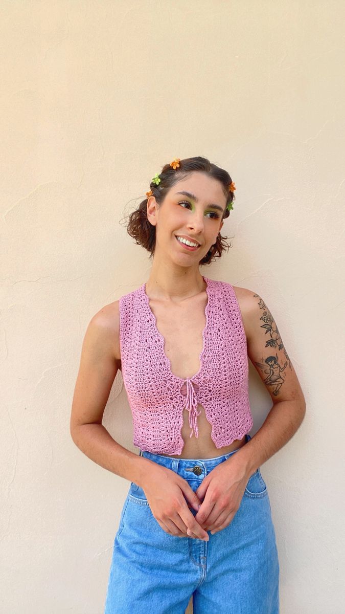 a woman with tattoos standing in front of a wall wearing blue jeans and a pink crop top