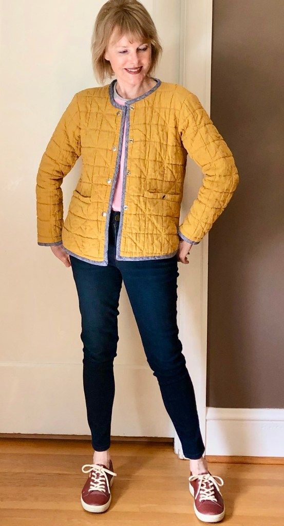 a woman standing in front of a wall wearing a yellow jacket and blue jeans with her hands on her hips