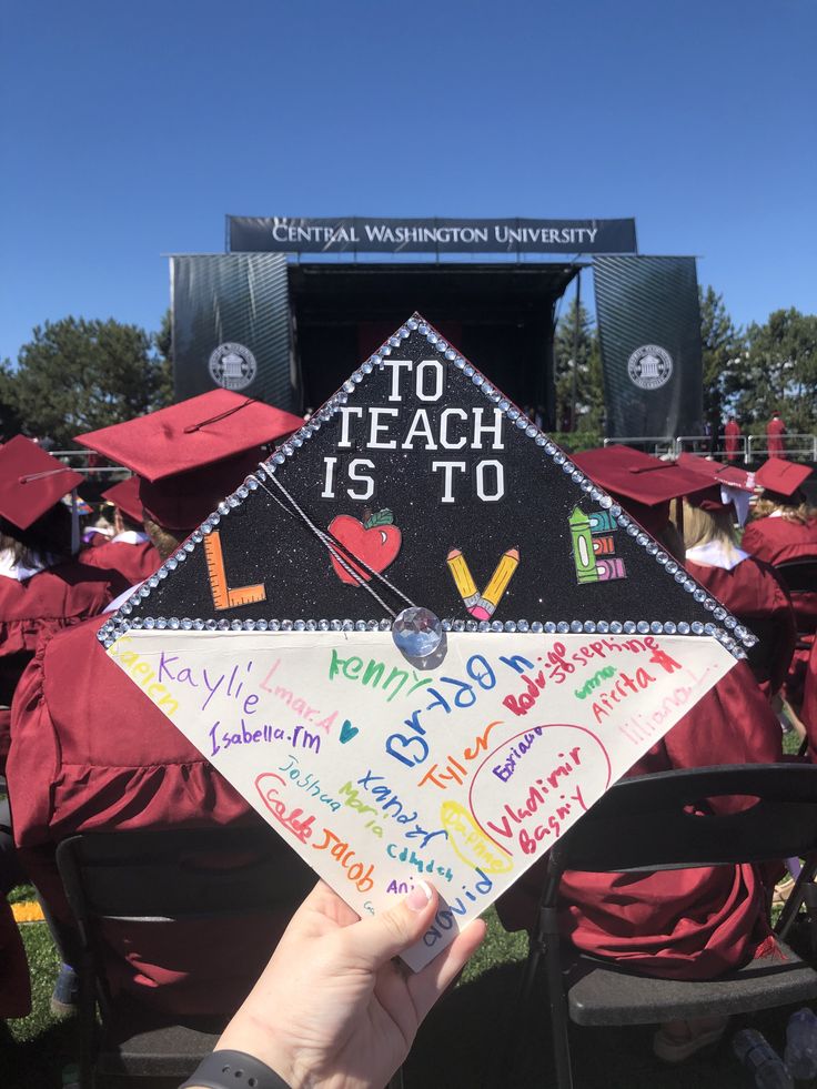 a hand holding up a sign that says to teach is to with other graduates in the background