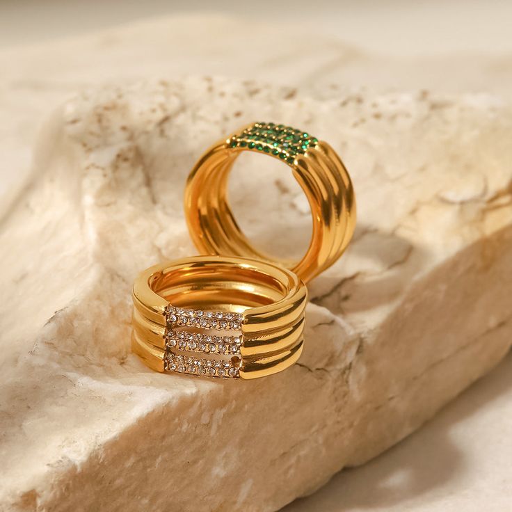 two gold rings with green and white diamonds on top of a rock next to each other