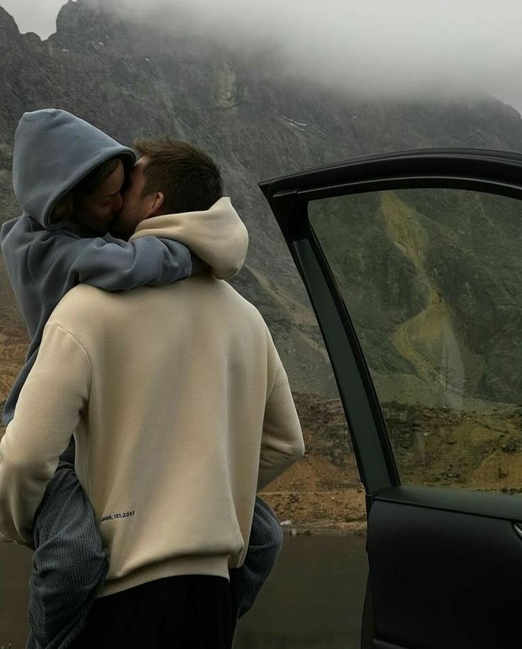 two people standing next to each other near a car with mountains in the back ground