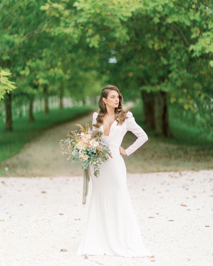 a woman in a white dress holding a bouquet standing on a dirt road surrounded by trees