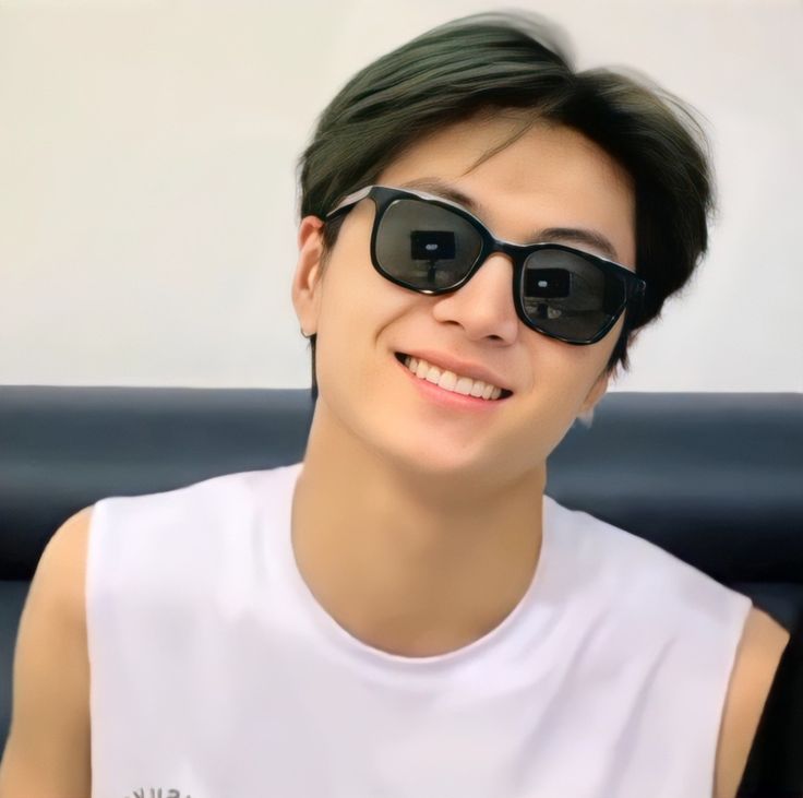 a young man wearing sunglasses and smiling at the camera while sitting on a couch in front of a white wall