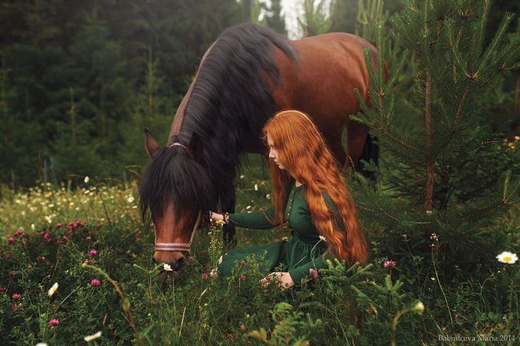 a woman with long red hair sitting in the grass next to a brown and black horse