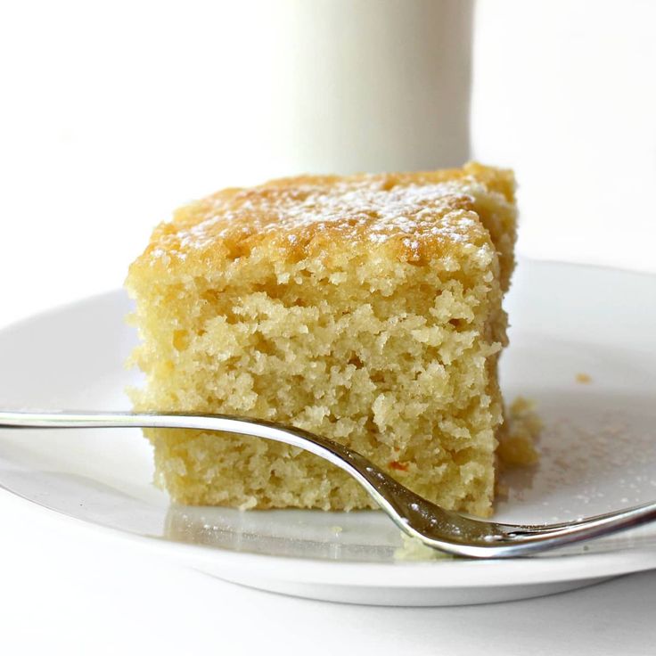 a piece of cake on a white plate with a fork next to it and a glass of milk in the background