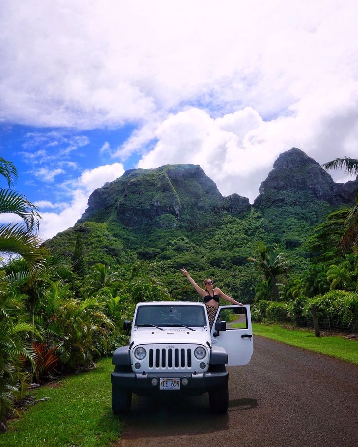 a woman standing in the back of a white jeep on a road next to lush green mountains