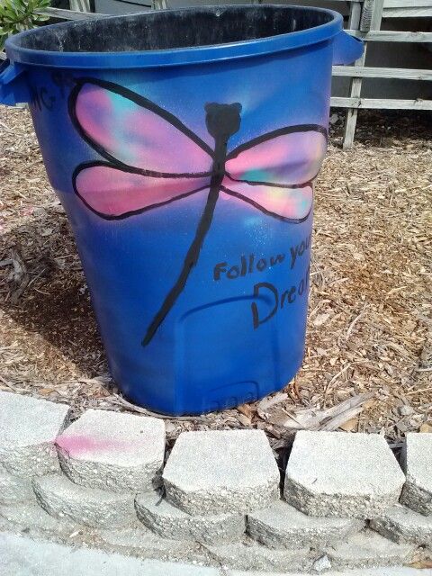 a blue bucket with a dragonfly painted on it sitting in front of a brick walkway