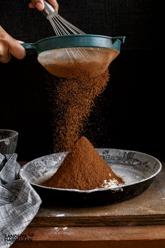 a person scooping brown powder from a bowl into a metal pan on top of a wooden table