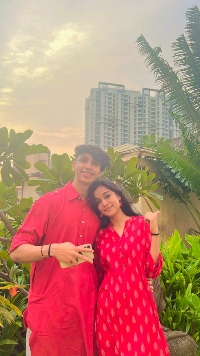 a man and woman standing next to each other in front of some plants with buildings in the background